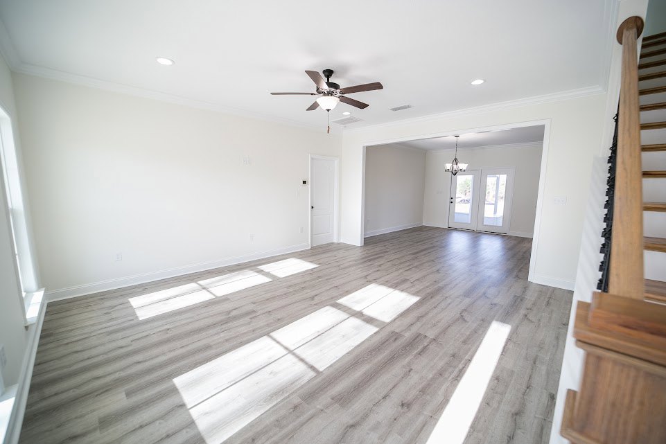 Ceiling fan with light fixture above wood flooring, white double doors with glass panes, white door with black knobs, sunlight streaming through windows onto floor, pale plaster