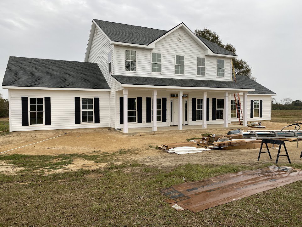 White house under construction with black shutters, pile of lumber on dirt, black sawhorse, brown plastic bag on grass, cloudy sky overhead