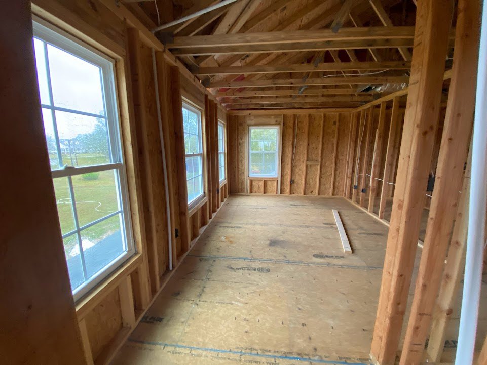 Living room with exposed wood ceiling beams, hardwood floors, large windows, and wood-paneled walls, overlooking grassy yard and neighboring house.