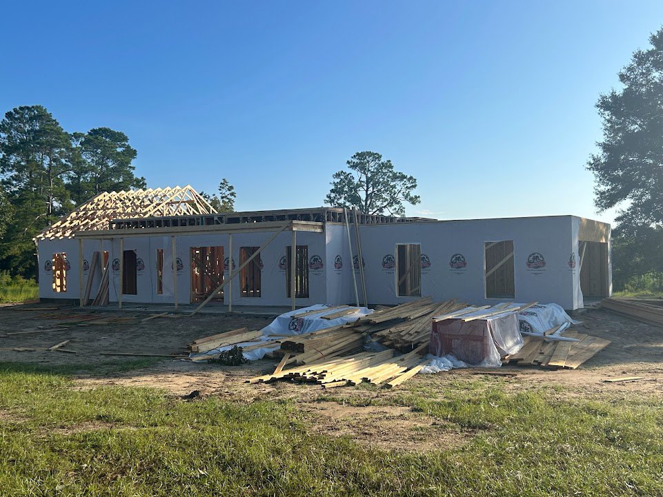 Wood-framed roof under construction, people working atop, man standing beside unfinished exterior, plastic-wrapped pallets on ground, white-trimmed window, grassy lot bordered by