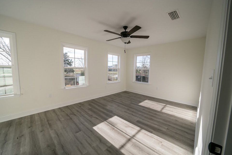 Bright room with wood laminate flooring, white plaster walls, ceiling fan with light fixture, and multiple windows showing views of trees, road, and nearby building.