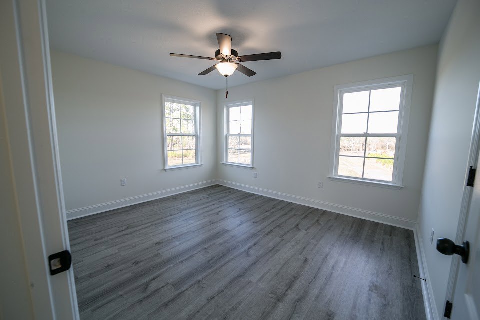 Ceiling fan with light fixture centered above wood laminate flooring, white framed windows along plaster walls, natural light highlighting interior finishes.