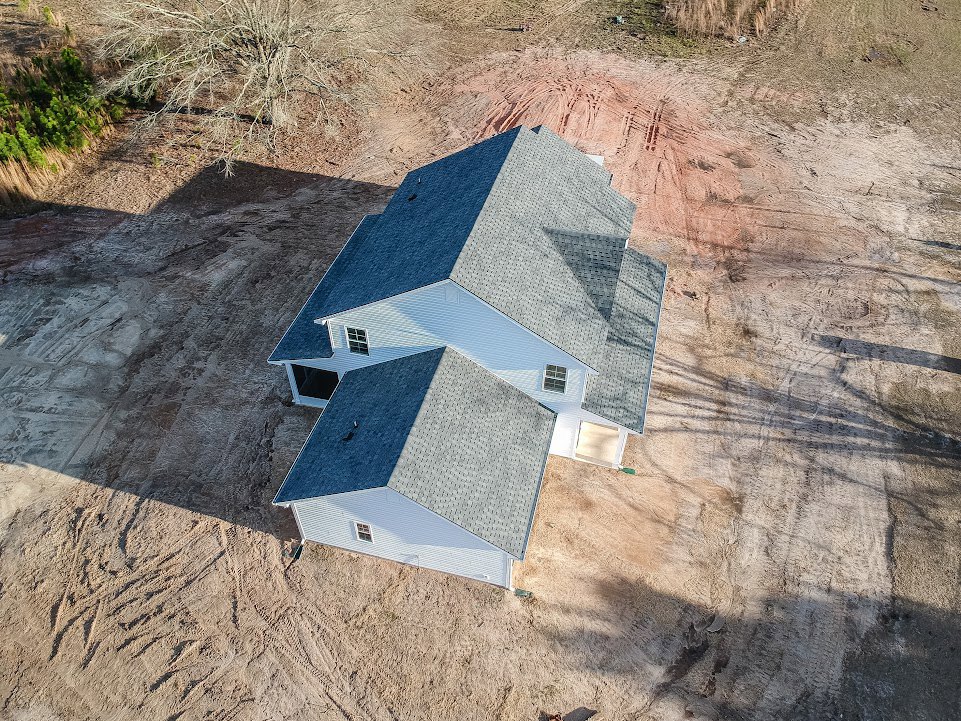 Blue-roofed house with large driveway, surrounded by bare tree, green bush, and expansive dirt yard