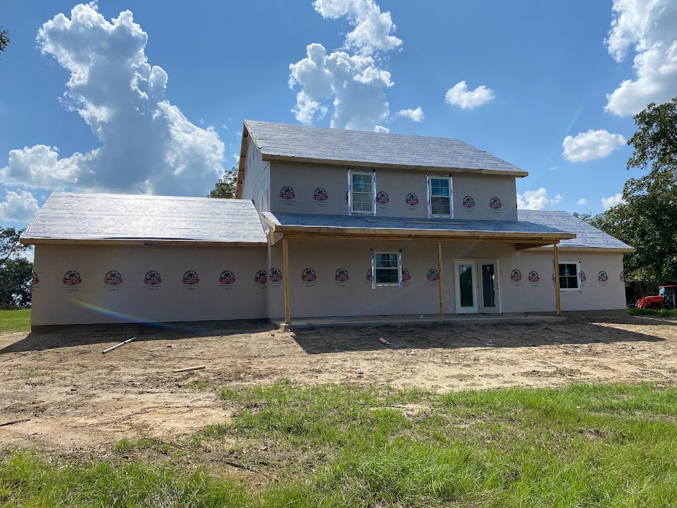Two-story house under construction with white-framed windows, glass-paneled door, exposed dirt yard, and grassy patches under a partly cloudy blue sky