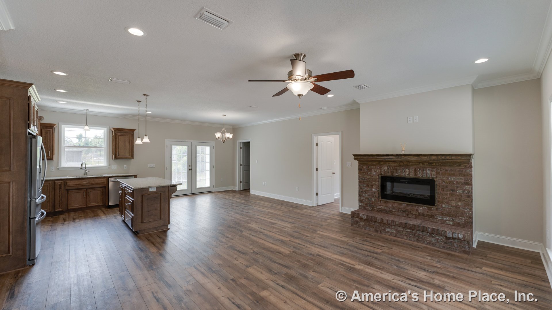 Open concept living area with wood-look flooring, brick fireplace with wood mantel, kitchen island topped with granite, stained wood cabinetry, double glass doors with built-in