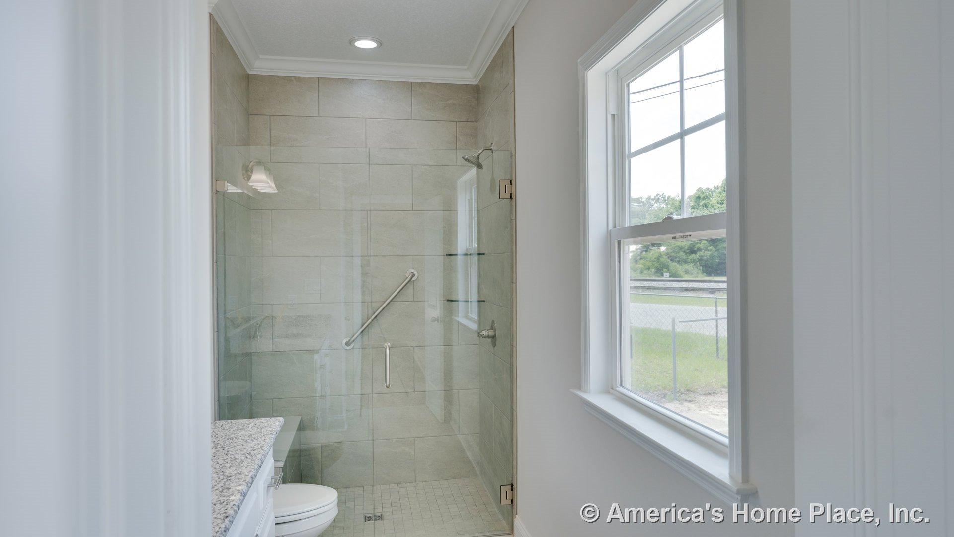 Walk-in shower with glass enclosure, large beige tile walls, built-in shower shelves, granite countertop vanity, white trim window, recessed ceiling light, and built-in grab bar.