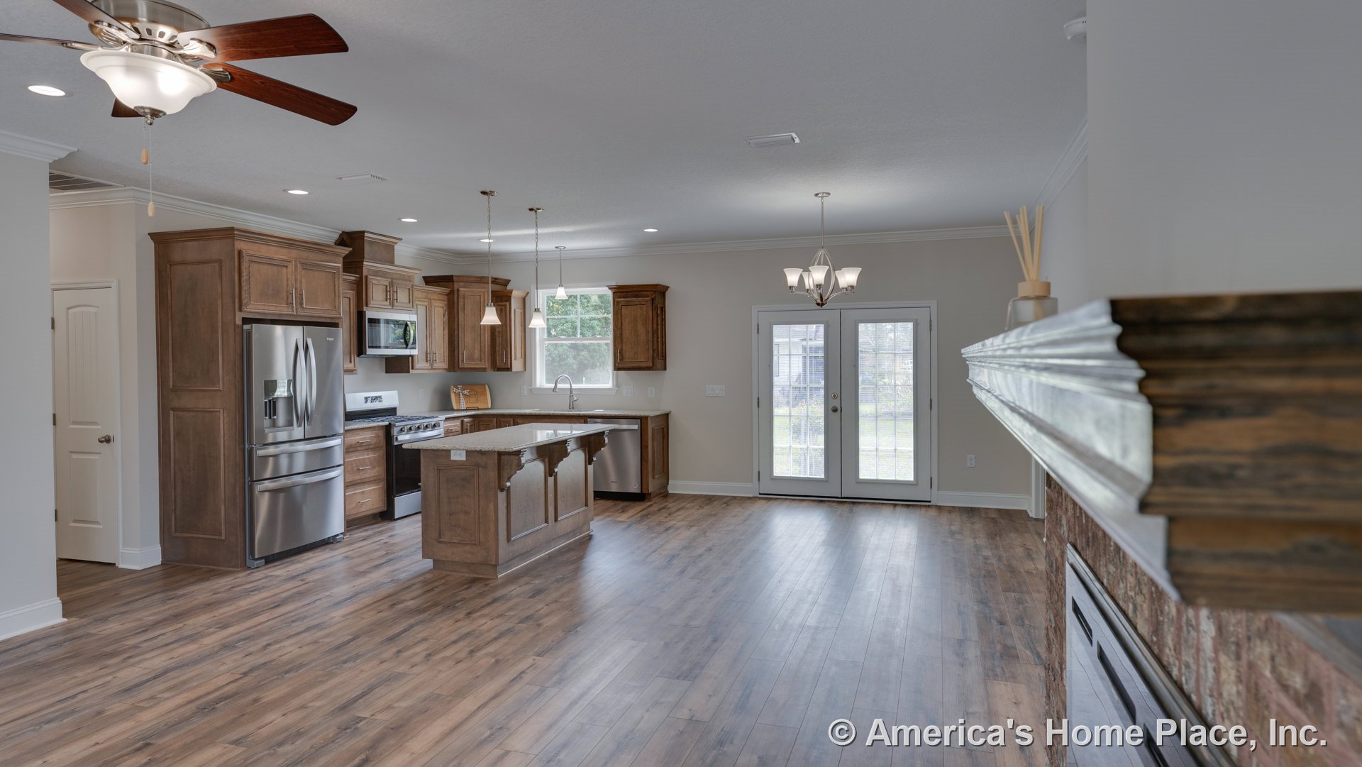 Open kitchen and living area with wood cabinetry, stainless steel refrigerator, center island with decorative corbels, brick fireplace topped by wood mantel, French doors with