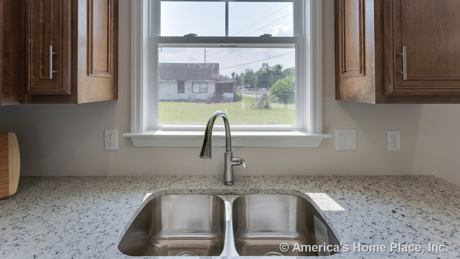 Double stainless steel sink with modern faucet set in granite countertop beneath a large window with trim, surrounded by wooden upper cabinets and electrical outlets on tiled