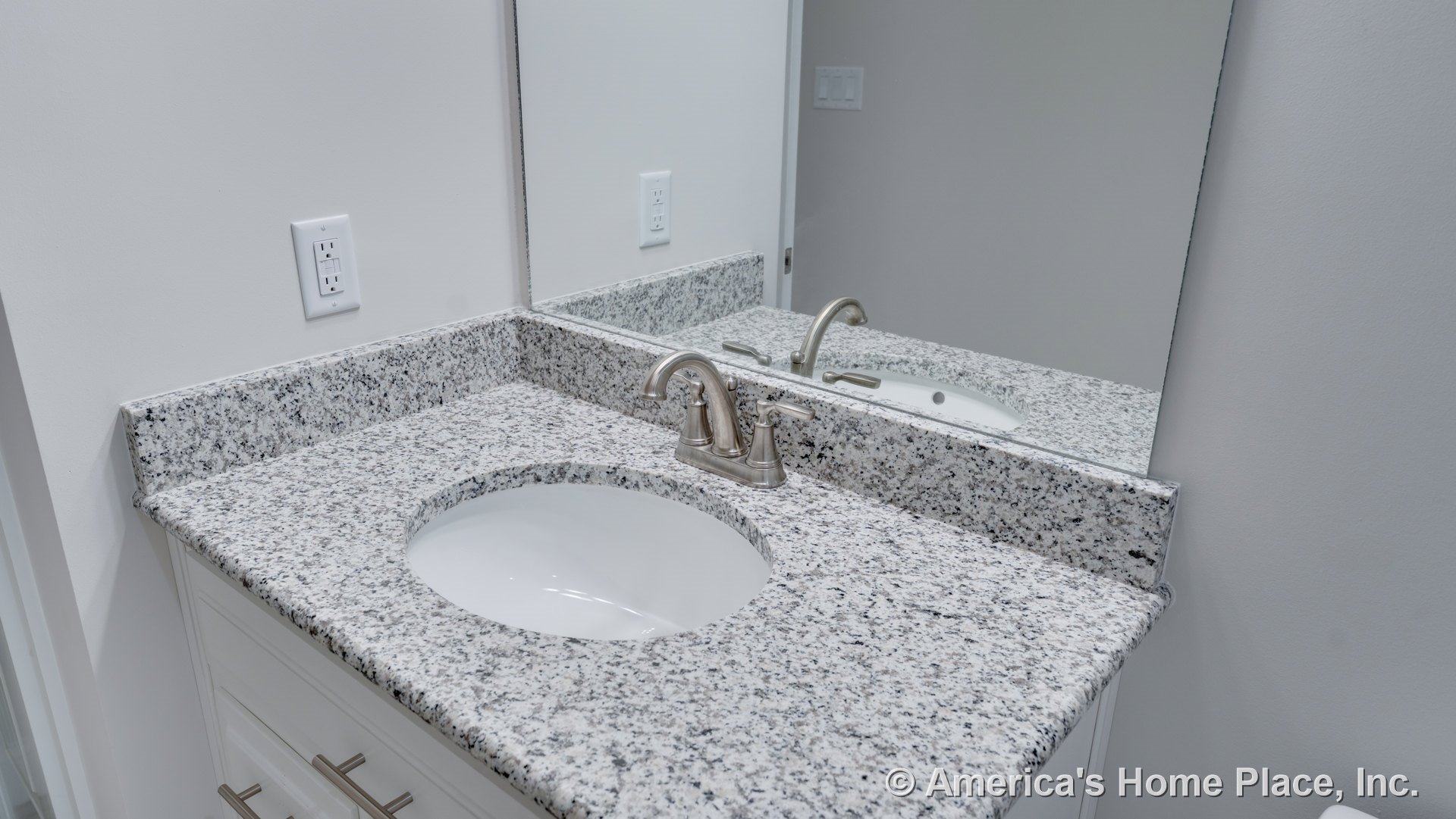 Speckled granite bathroom vanity with undermount sink, brushed nickel faucet, large wall mirror, white cabinetry, and wall outlet.
