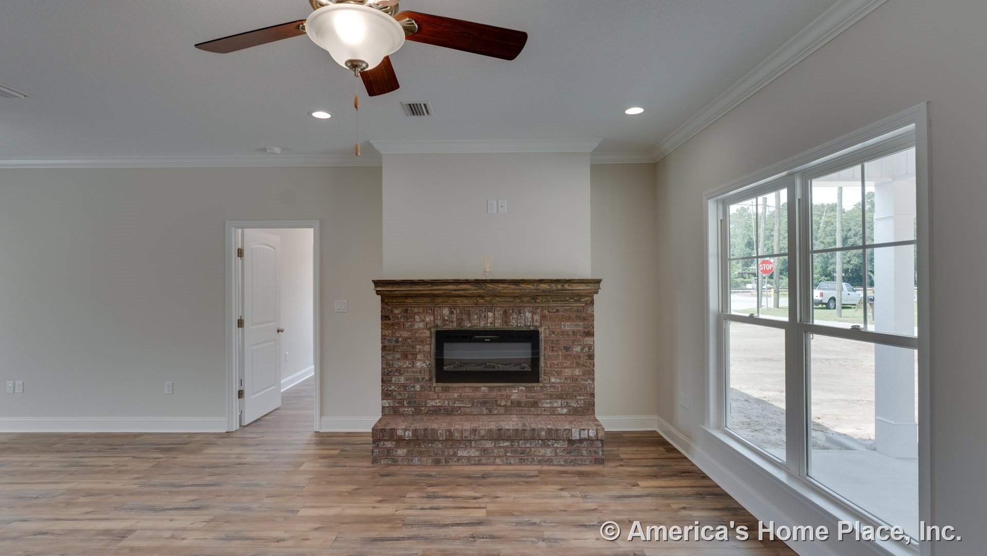 Brick fireplace with wood mantel centered between large double windows, ceiling fan with light overhead, wood-look flooring, white trim and crown molding, recessed lighting, and