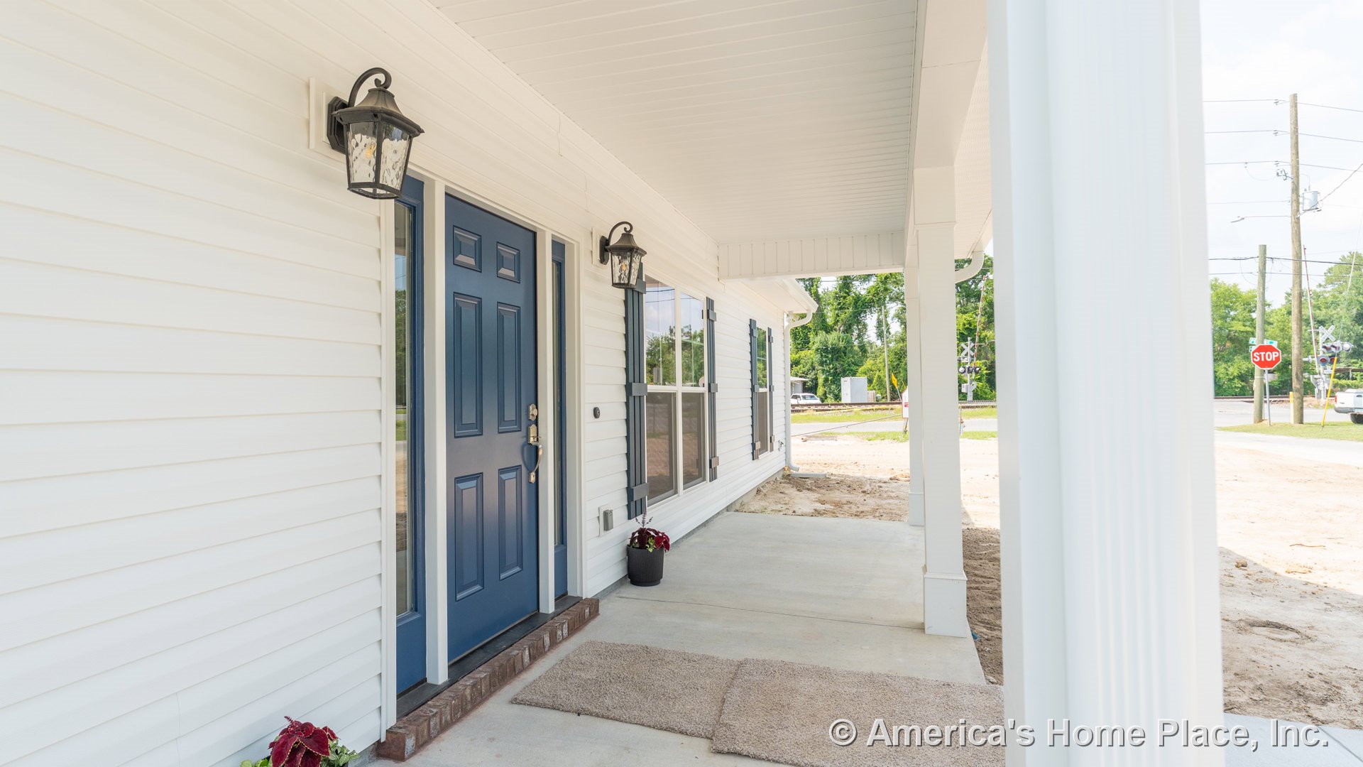 Blue paneled entry door set in white vinyl siding, covered porch ceiling with outdoor lantern fixtures, concrete porch floor, windows with shutters, decorative trim, and supporting