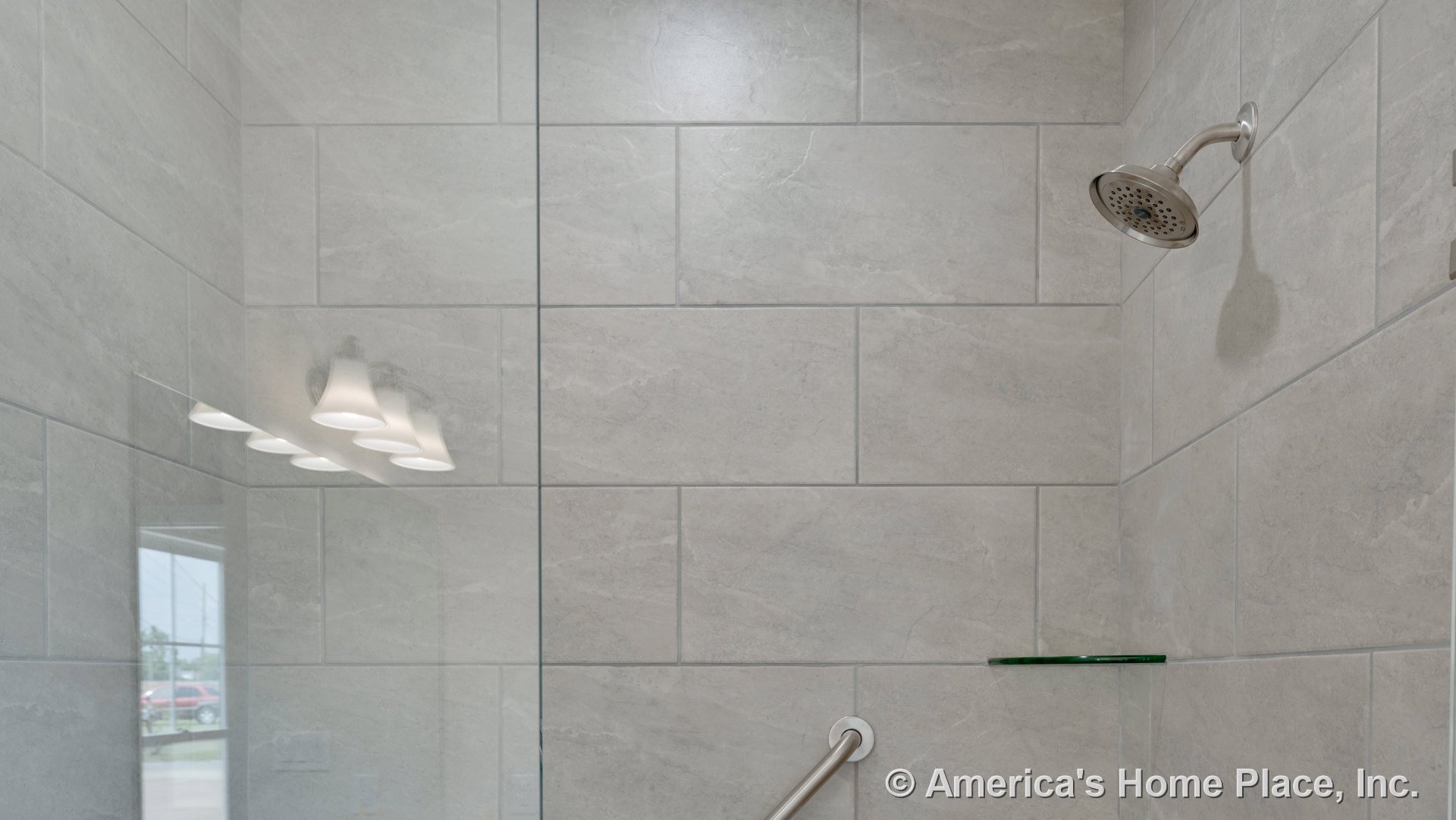 Large rectangular gray tile shower walls with stainless steel showerhead, built-in glass shelf, grab bar, and reflected ceiling light in a modern bathroom.