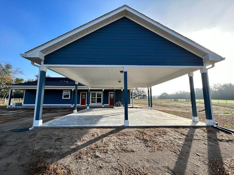 Blue siding home with white pillars supporting a covered front porch, surrounded by trees and landscaped yard under a clear sky