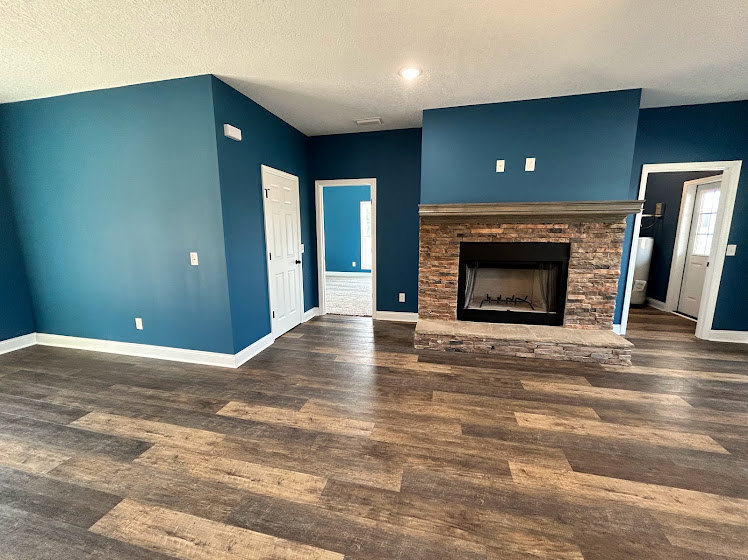 Living room featuring a stone fireplace, wide plank hardwood floors, neutral walls, and large windows with natural light.