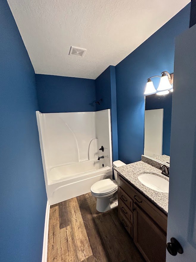 Bathroom with freestanding white bathtub, white sink with black faucet, toilet, wood flooring, white baseboards, and light-colored tile walls.