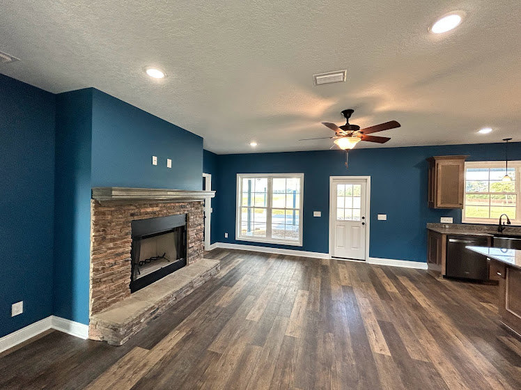 Living room featuring wood flooring, stone fireplace with white mantel, neutral walls, ceiling fan, and built-in cabinetry
