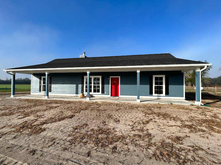 Blue siding exterior with white trim, red front door, covered porch, manicured lawn, and partly cloudy sky