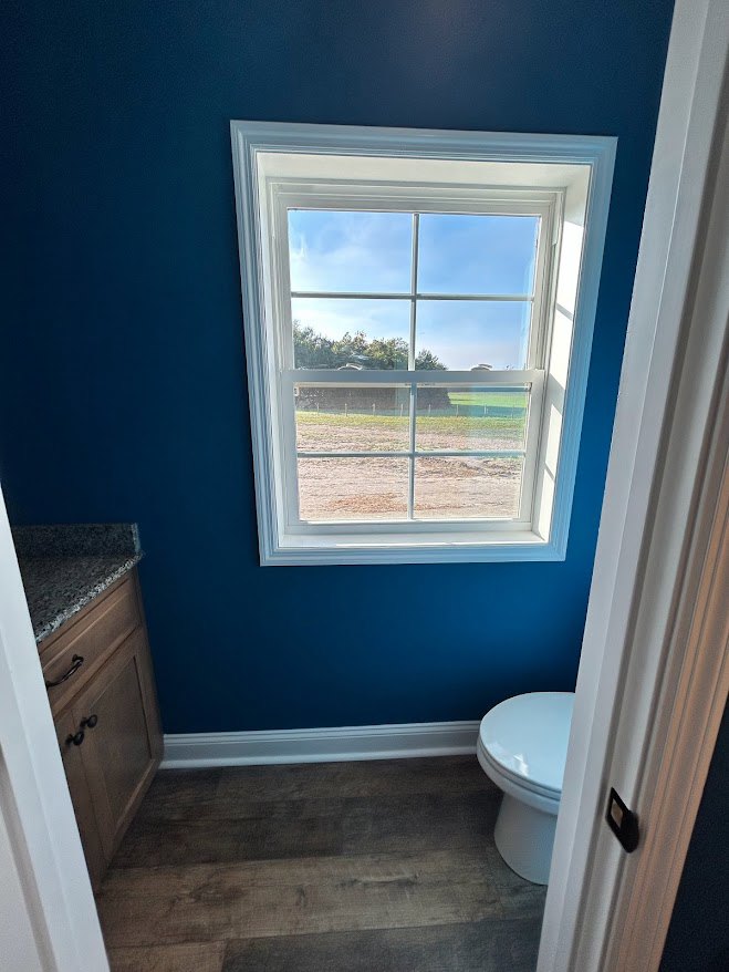 Bathroom with wood flooring and white trim, toilet beside a window overlooking a green field under blue sky with clouds