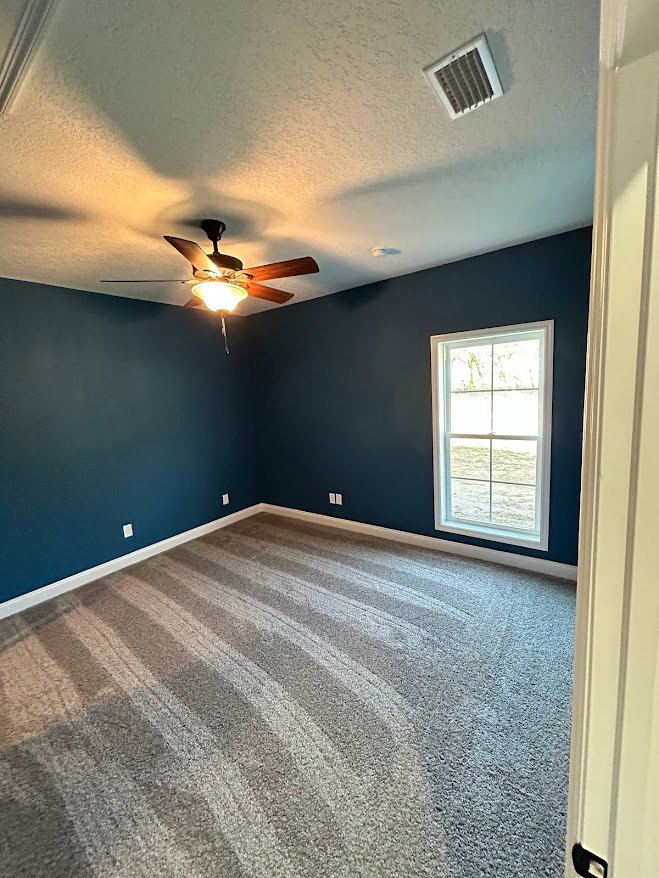 Carpeted room with white walls, ceiling fan and light fixture, window with white frame, ceiling vent, and white baseboards