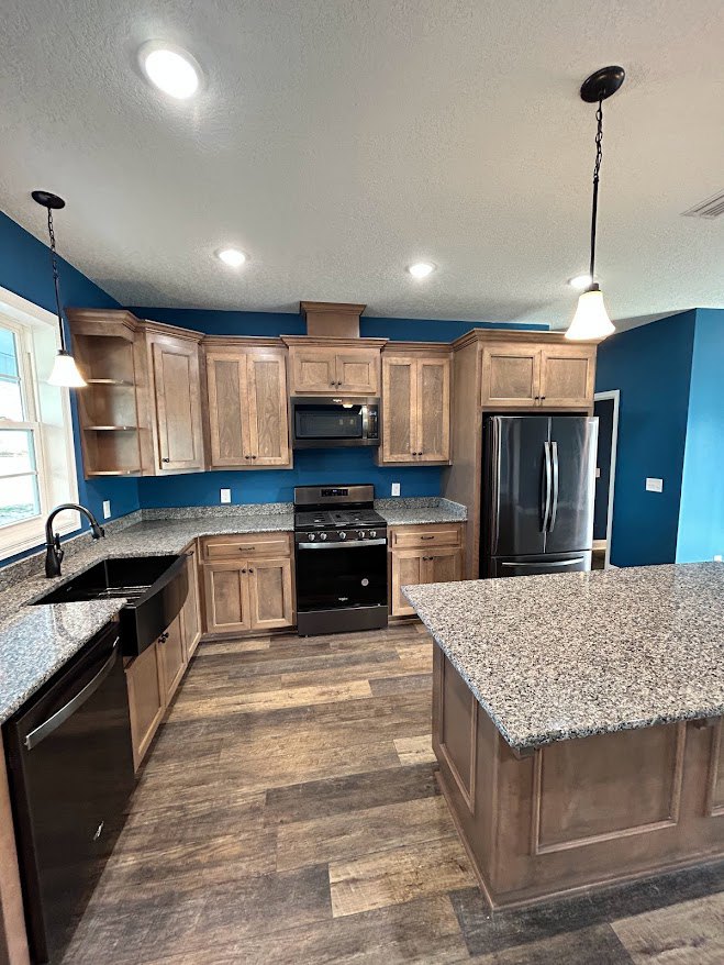 Granite countertop kitchen with wood base cabinets, black refrigerator, stove, dishwasher, stainless steel sink, and overhead lamp.