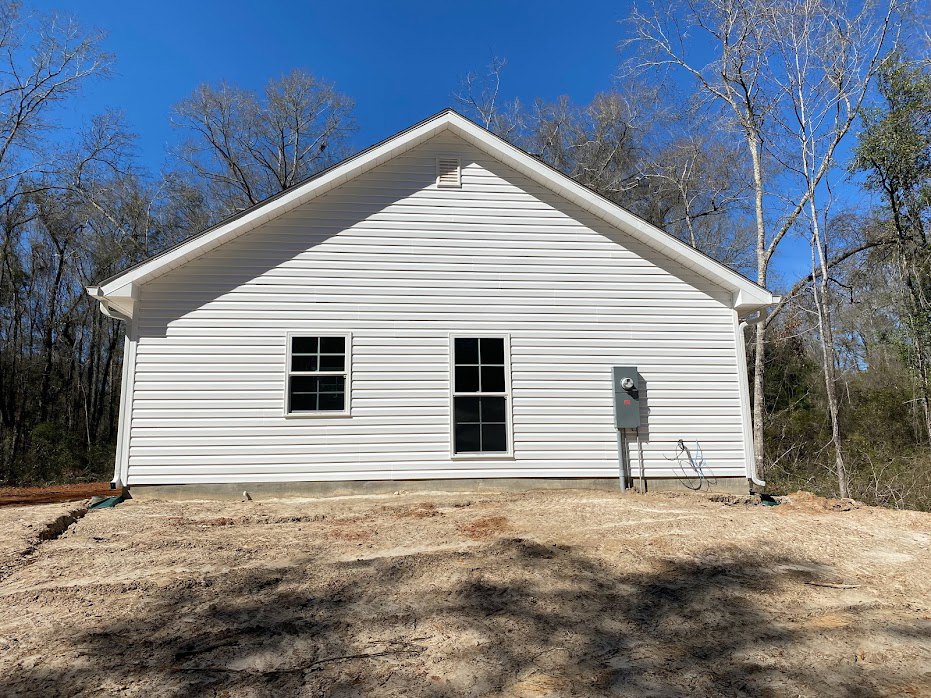 White house with horizontal siding, rectangular side addition, white-framed windows, dirt yard, trees in background, blue sky overhead