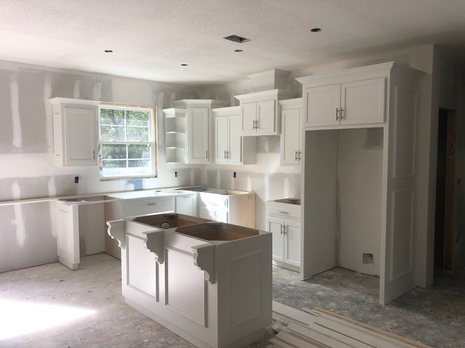 White kitchen with shaker cabinets, black handles, white island with drawers, tile backsplash, stainless sink under a window with white trim, light fixture illuminating quartz