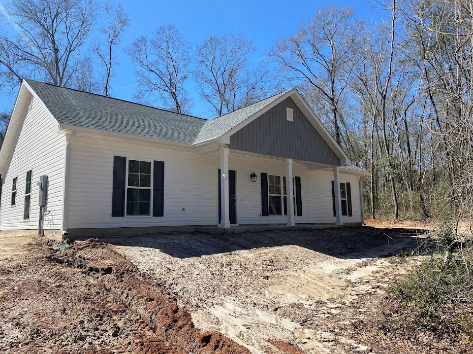 White house with black shutters and white pillars, dirt yard in foreground, trees in background, white-framed windows