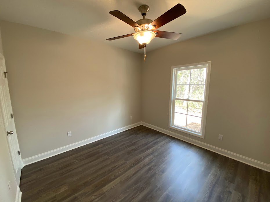 Ceiling fan with light fixture mounted on white ceiling, large window showing leafy trees, wood flooring, white door with silver doorknob, pale walls.