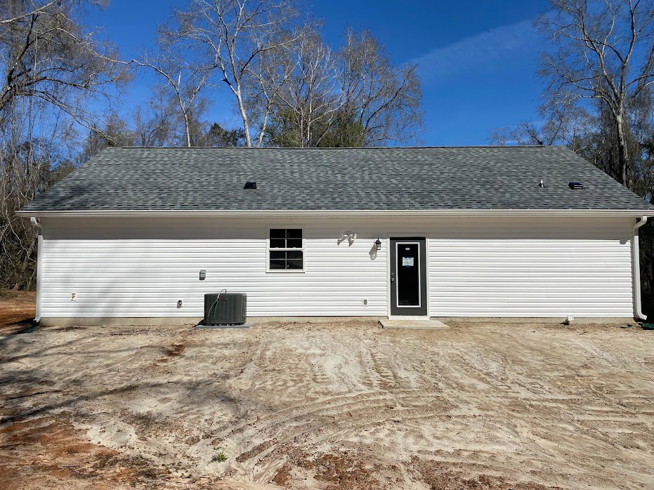White house with black door and white-framed window, dirt and tire tracks in front yard, black rectangular object with cord near entrance, cloudy sky and trees in background