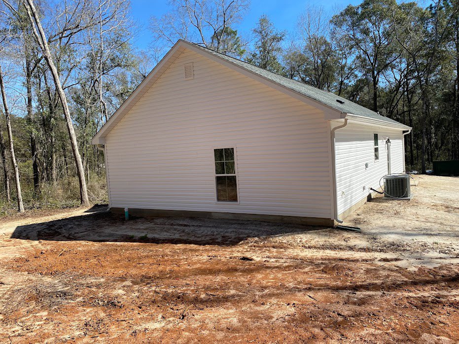 White house with white siding, garage door, white-framed windows, air conditioner unit, dirt yard, and trees in the background