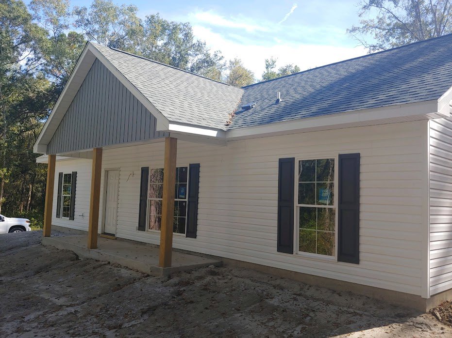 Two-story home with gray siding, covered front porch, concrete driveway, white-trimmed windows, and a dirt patch in the yard