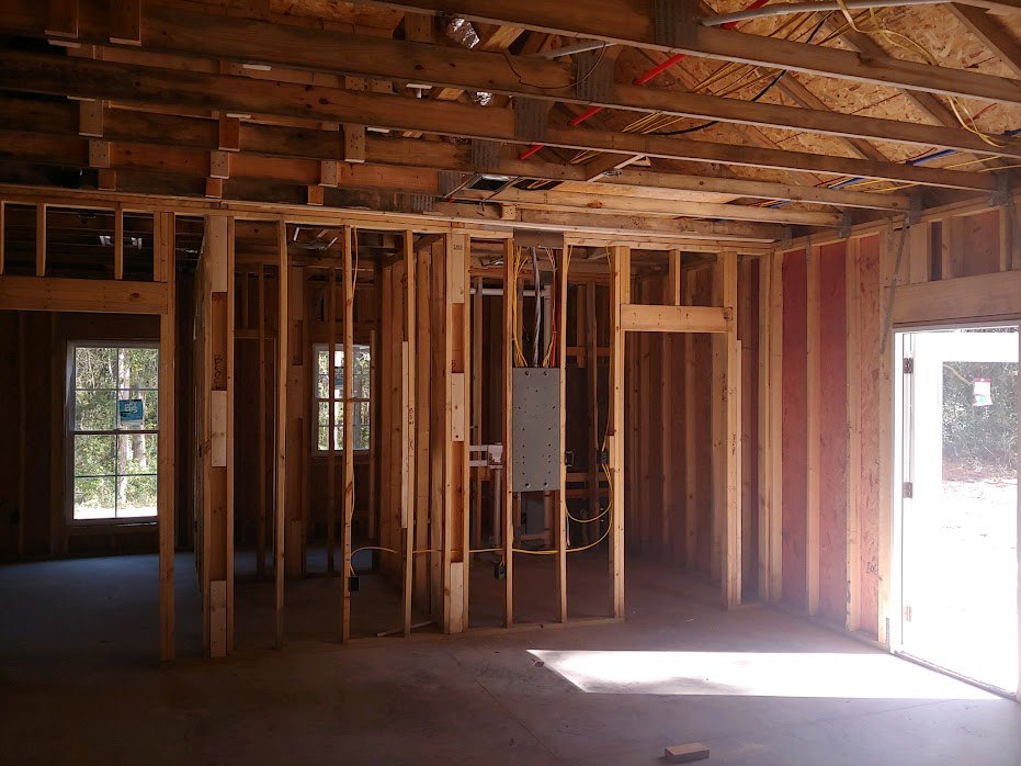 Exposed wood framing with ceiling beams, unfinished grey wall with black specks, window displaying a sign, adjacent white door, sunlight illuminating construction floor