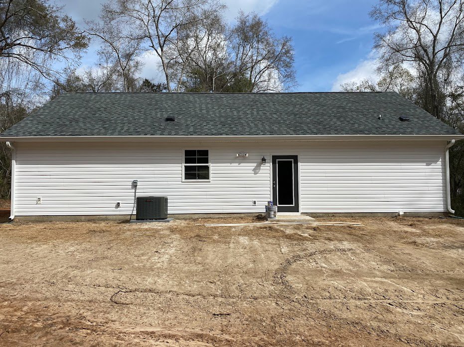 White cottage-style home with black front door and white-framed windows, surrounded by dirt lot and scattered trees under a partly cloudy sky