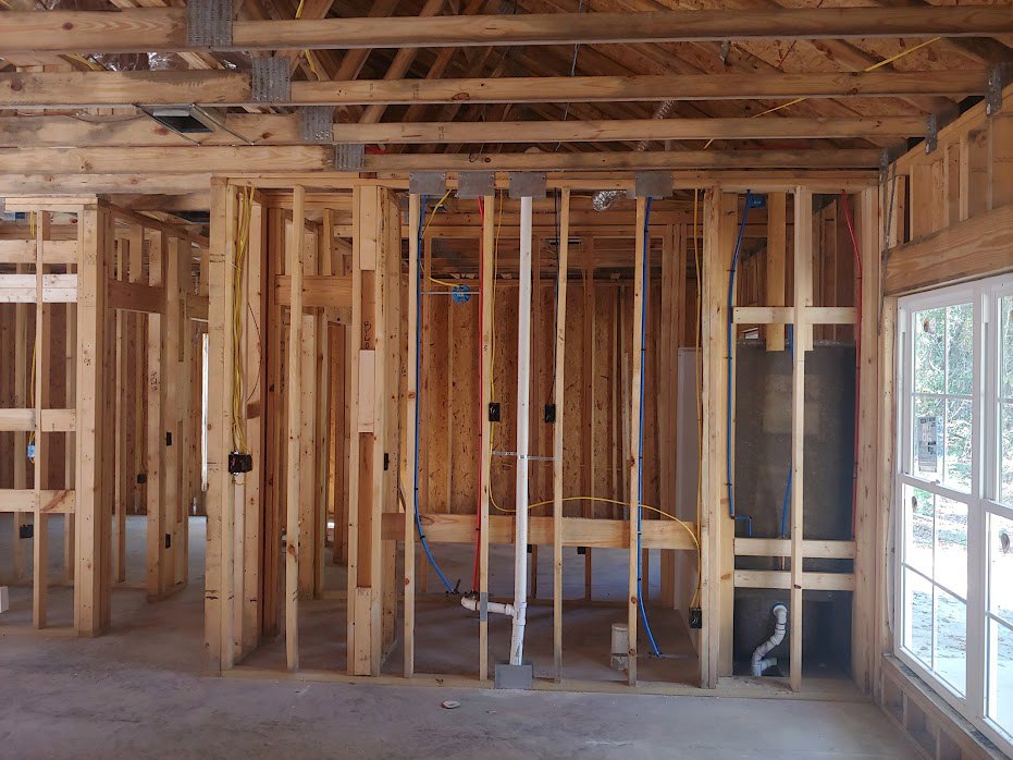 Exposed wood framing with ceiling beams, white door featuring glass panes, visible electrical wiring, blue plumbing pipes, and white pipe in unfinished interior space