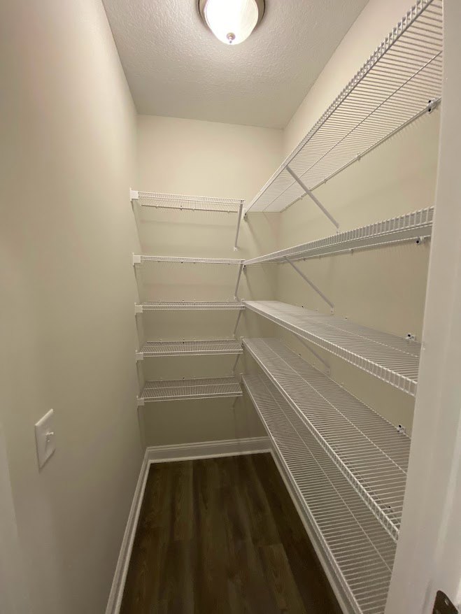 Empty closet with white shelves, dark wood floor with vent, recessed ceiling light, smooth plaster walls