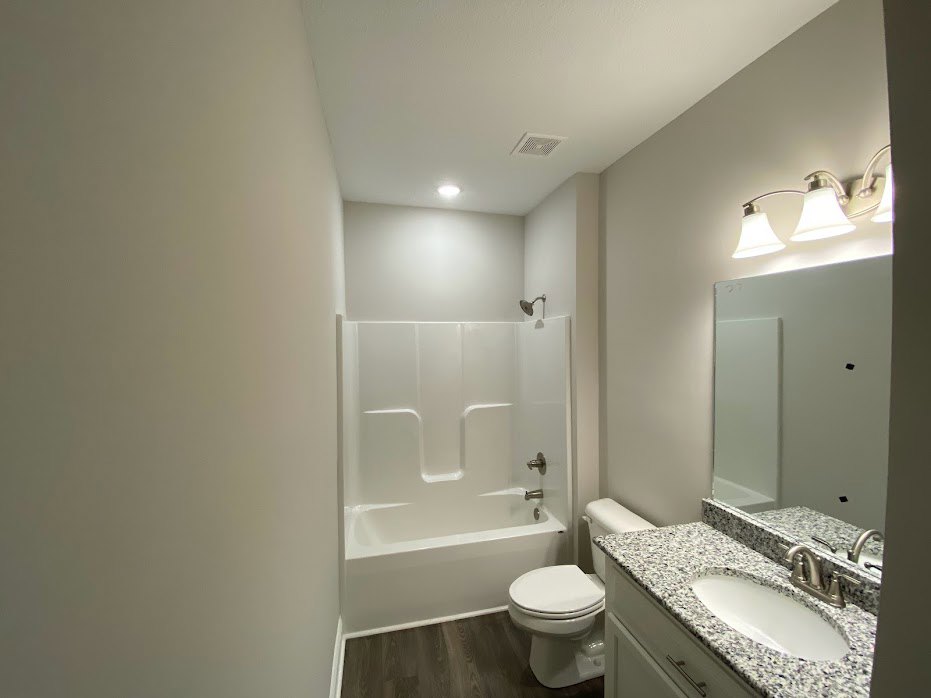 White porcelain sink set in a granite countertop, silver faucet, adjacent toilet and bathtub with silver handles, light fixture with two lamps mounted above, neutral tile walls and