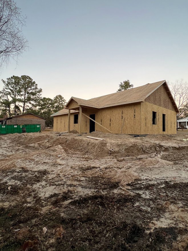 Wood-framed house under construction on a dirt lot with scattered debris, leafless tree nearby, green storage container, and bare trees in the background