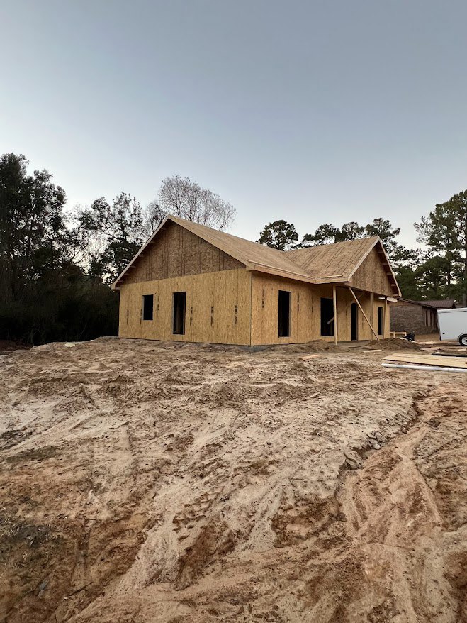 Wood-framed house under construction on a dirt lot, unfinished roof and walls, surrounded by sparse trees and open sky