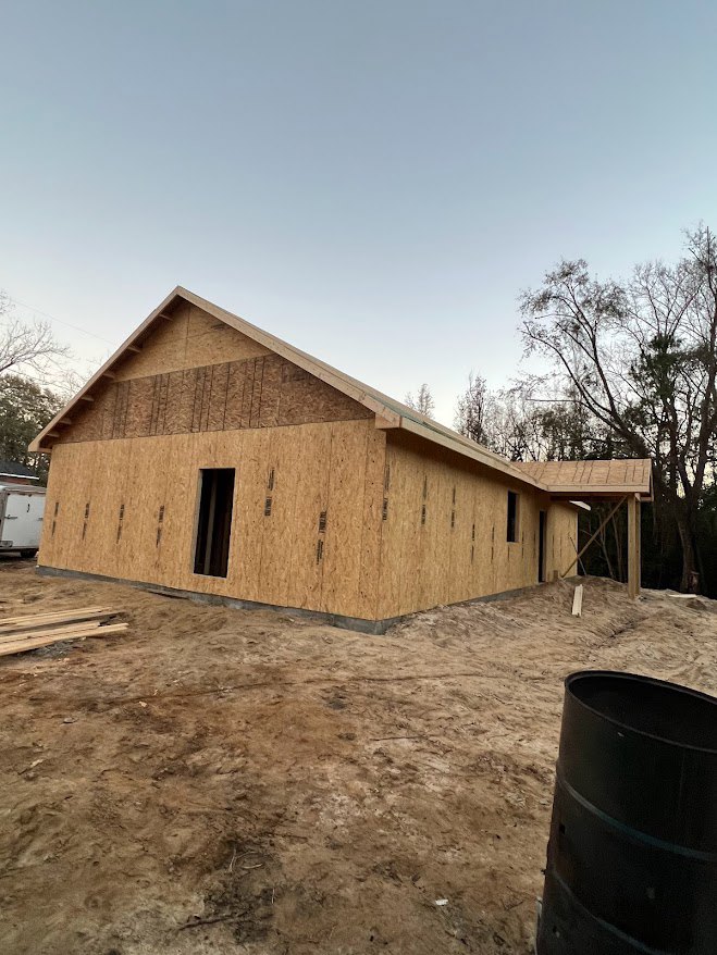 Framed custom home under construction with exposed wood, black barrel on dirt, pile of lumber, and trees in background