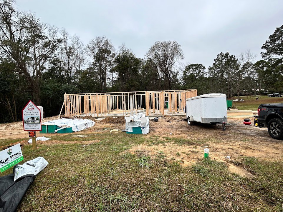 Wood-framed custom home under construction in grassy field, black truck and white trailer parked nearby, construction sign with house rendering in foreground, scattered building