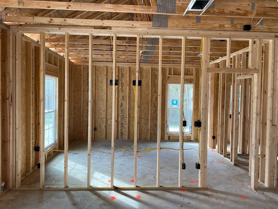 Wood-framed room under construction with exposed beams, white-framed windows, unfinished wooden planks, and a blue sign taped to one window