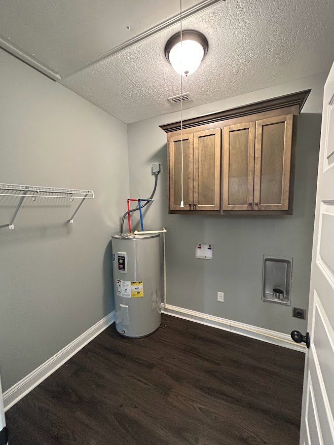 Utility room with a large white water tank featuring a yellow label, dark wood flooring, white baseboards, and white cabinetry along the wall
