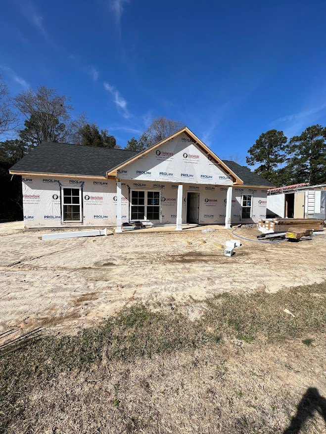 Partially built house with white sheathing, exposed window with permit sign, dirt yard in foreground, pitched roof, surrounding trees, and blue sky overhead