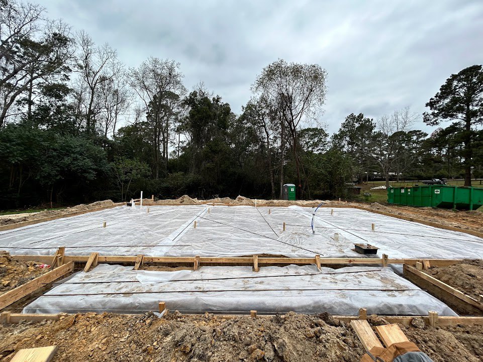 Concrete foundation under construction with plastic sheeting, green dumpster with white sticker, pile of dirt and rocks, surrounding trees and bushes, cloudy sky overhead
