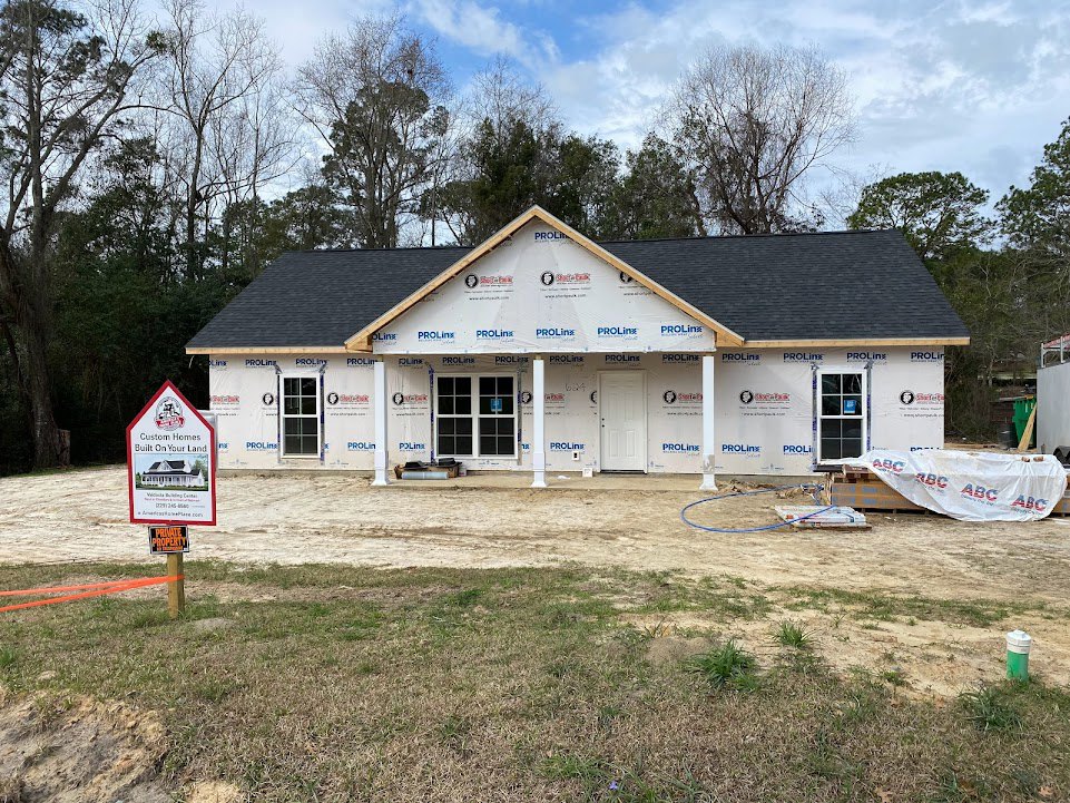Two-story house under construction with white roof, white plastic wrap with red and blue writing, black-trimmed white door, and a yard sign displaying a house illustration.
