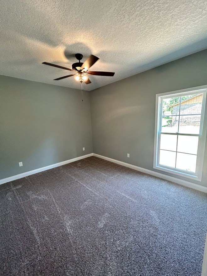 Bedroom with beige carpet flooring, white walls, ceiling fan with light fixture, and large window framed in white trim