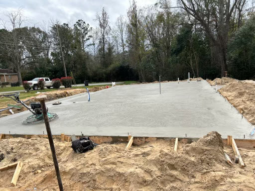 Freshly poured concrete slab foundation with construction equipment, surrounded by soil, trees, and open sky