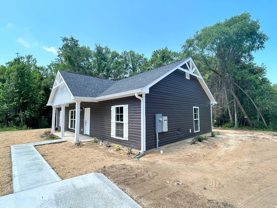 Framed house under construction with white door and window, white trim, concrete walkway beside dirt patch, trees and fallen branches in background, small white cottage visible