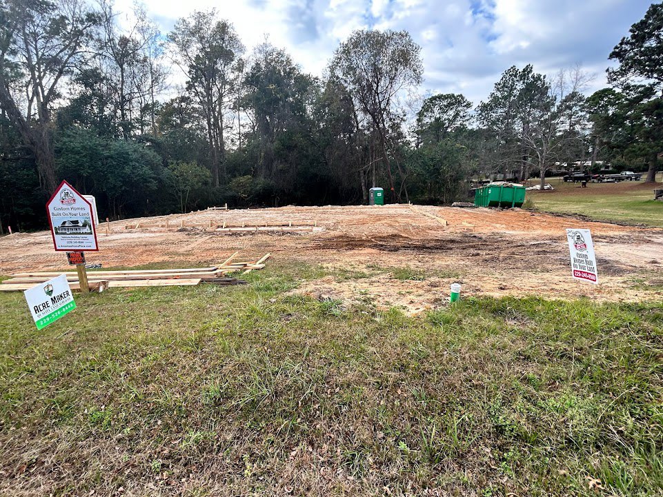 Grassy construction site with dirt path, pile of wood, house illustration sign, scattered trees, blue sky, and clouds
