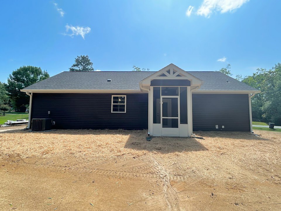 Modern house exterior with covered porch, white door featuring glass panels, large window with white frame, tire tracks visible in dirt driveway, surrounding trees and cloudy sky